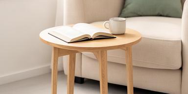 Mesa auxiliar redonda de madera clara junto a sillón con libro y taza en departamento compacto bien iluminado